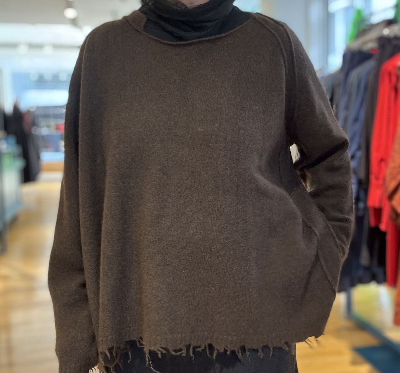 Person trägt oversized olivfarbenen Turtleneck-Pullover mit Fransen in einem Geschäft.