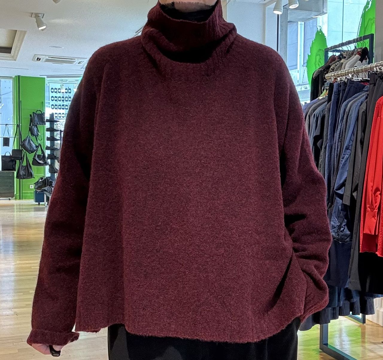 Person trägt burgunderfarbenen oversized Pullover mit schwarzen Hosen in einem Geschäft.