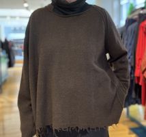 Person trägt oversized olivfarbenen Turtleneck-Pullover mit Fransen in einem Geschäft.