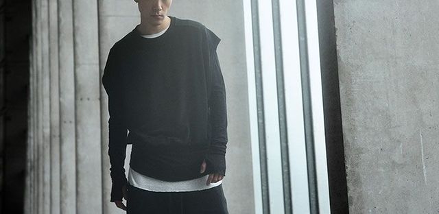 Mann in schwarzem Hoodie und hellem T-Shirt lehnt lässig an Säulen.