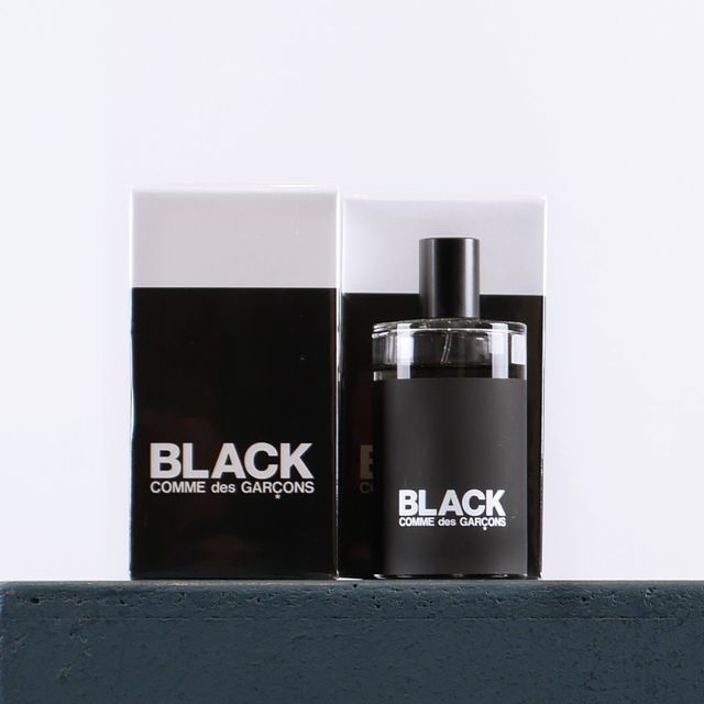 Schwarzes Parfüm „Black Comme des Garçons“ mit Flakon und Karton auf grauer Fläche.