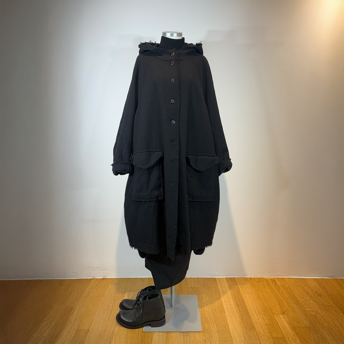 Langer, dunkler Wollmantel mit Pelzkragen und Taschen auf Mannequin, kombiniert mit schwarzen Stiefeln.