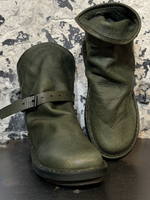 Alt-Text: Abgetragene grüne Stiefel auf Metallplatte vor schmutziger Wand.