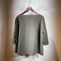 Graue, oversized Langarm-Bluse hängt an Holzklamotte mit kariertem Stoff.