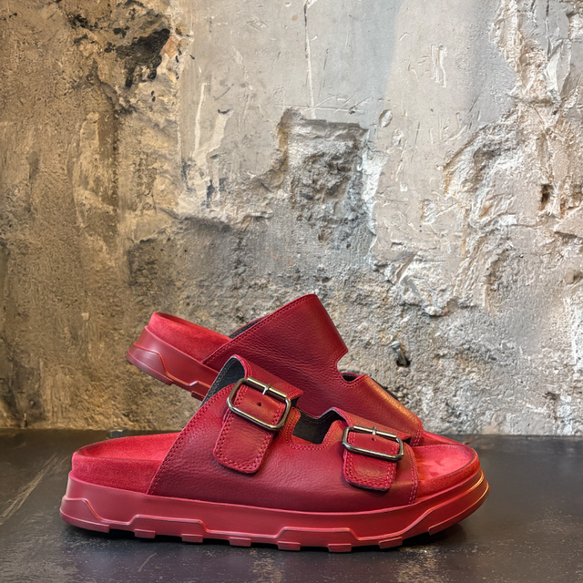 Rote Ledersandalen mit Buckelverschluss auf dunklem Untergrund vor texturierter Wand.